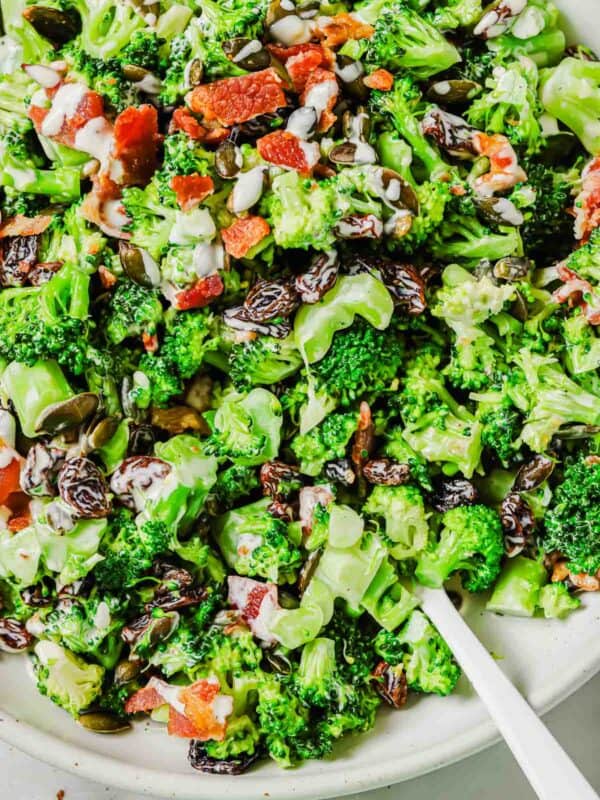 A close-up of a broccoli raisin salad mixed with bacon bits and seeds served on a white plate with a white utensil visible to the side.